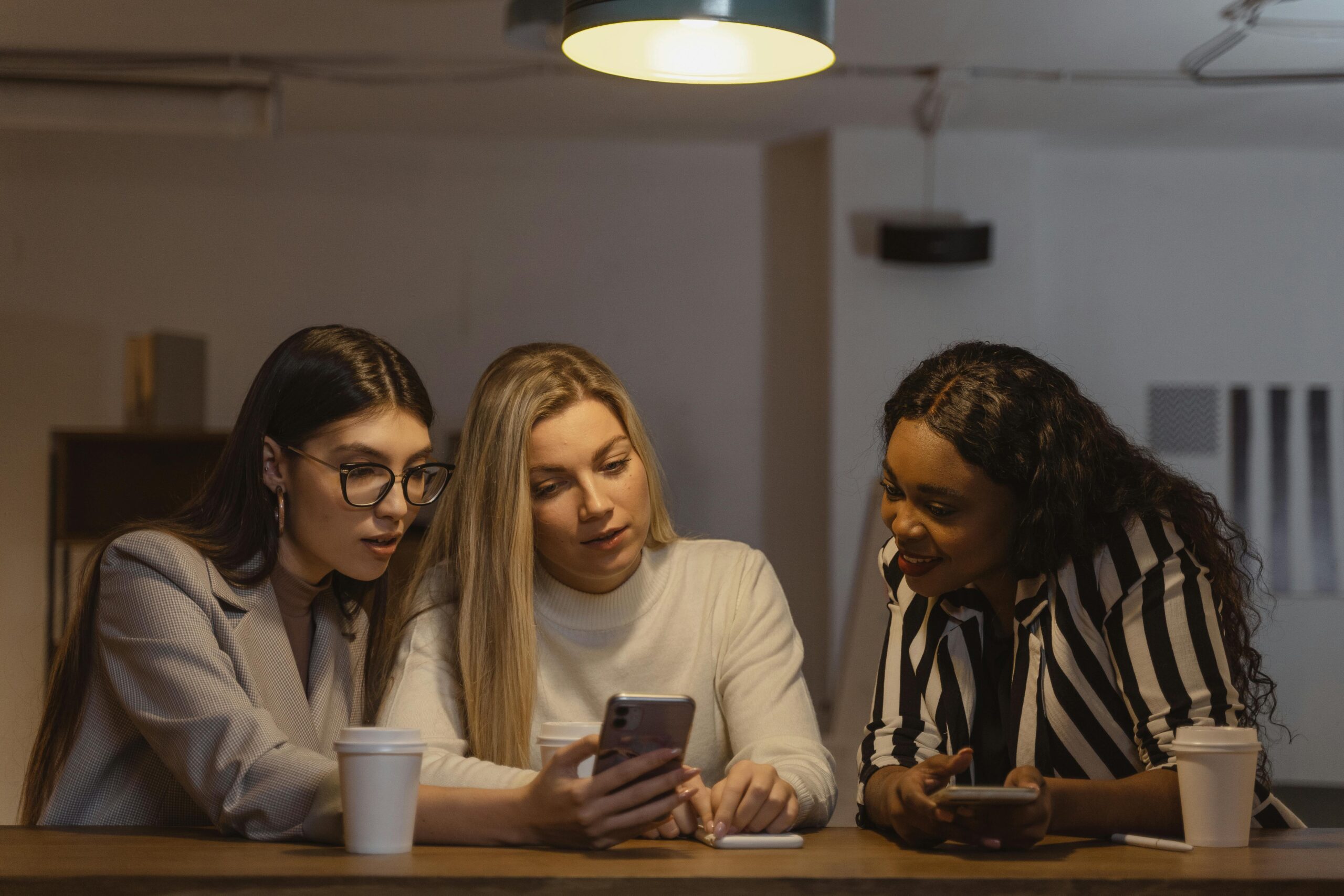 Three diverse women enjoying time together, interacting with smartphones at a table indoors.